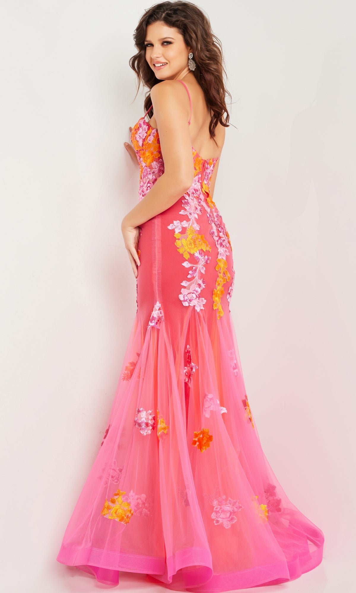 A woman wears the Long Prom Dress 36843 by Jovani, a pink mermaid tulle gown featuring yellow, orange, and white floral embroidery, as she stands against a plain light background and glances over her shoulder.